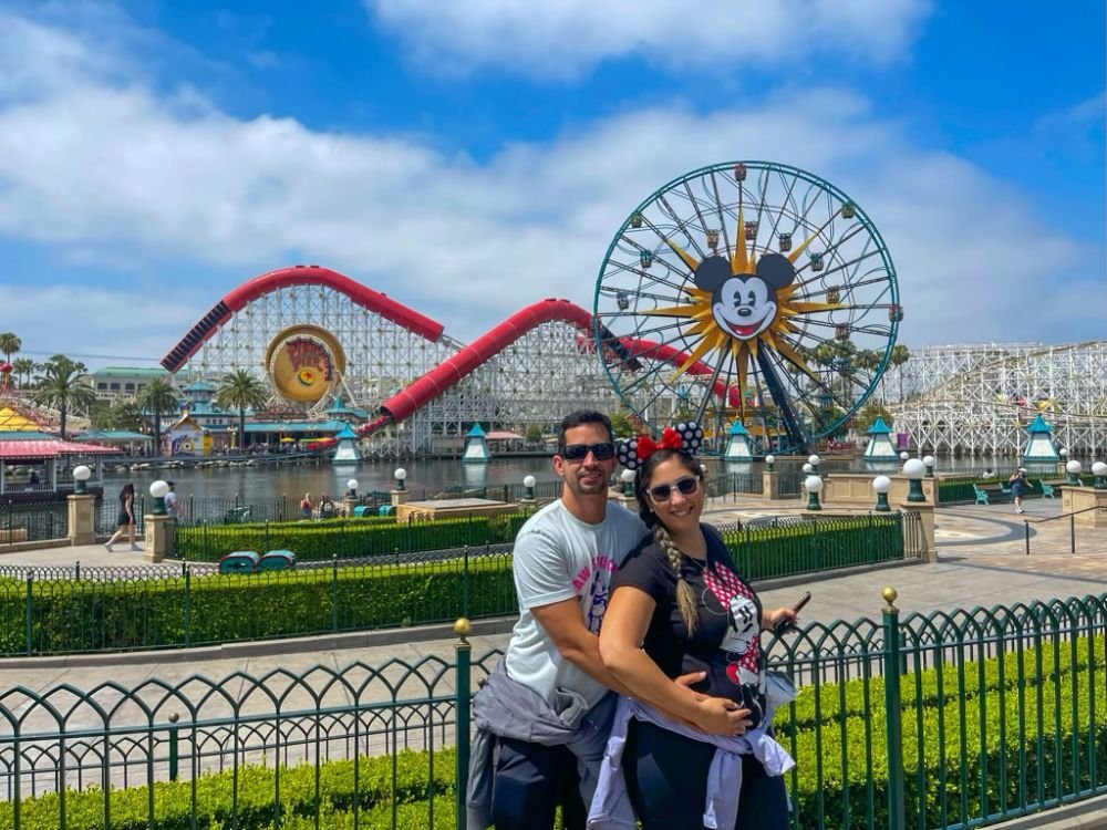 Foto do post Disney California Adventure com Crianças Pequenas: Casal com a imagem tradicional da roda gogante e montanha-russa ao fundo.